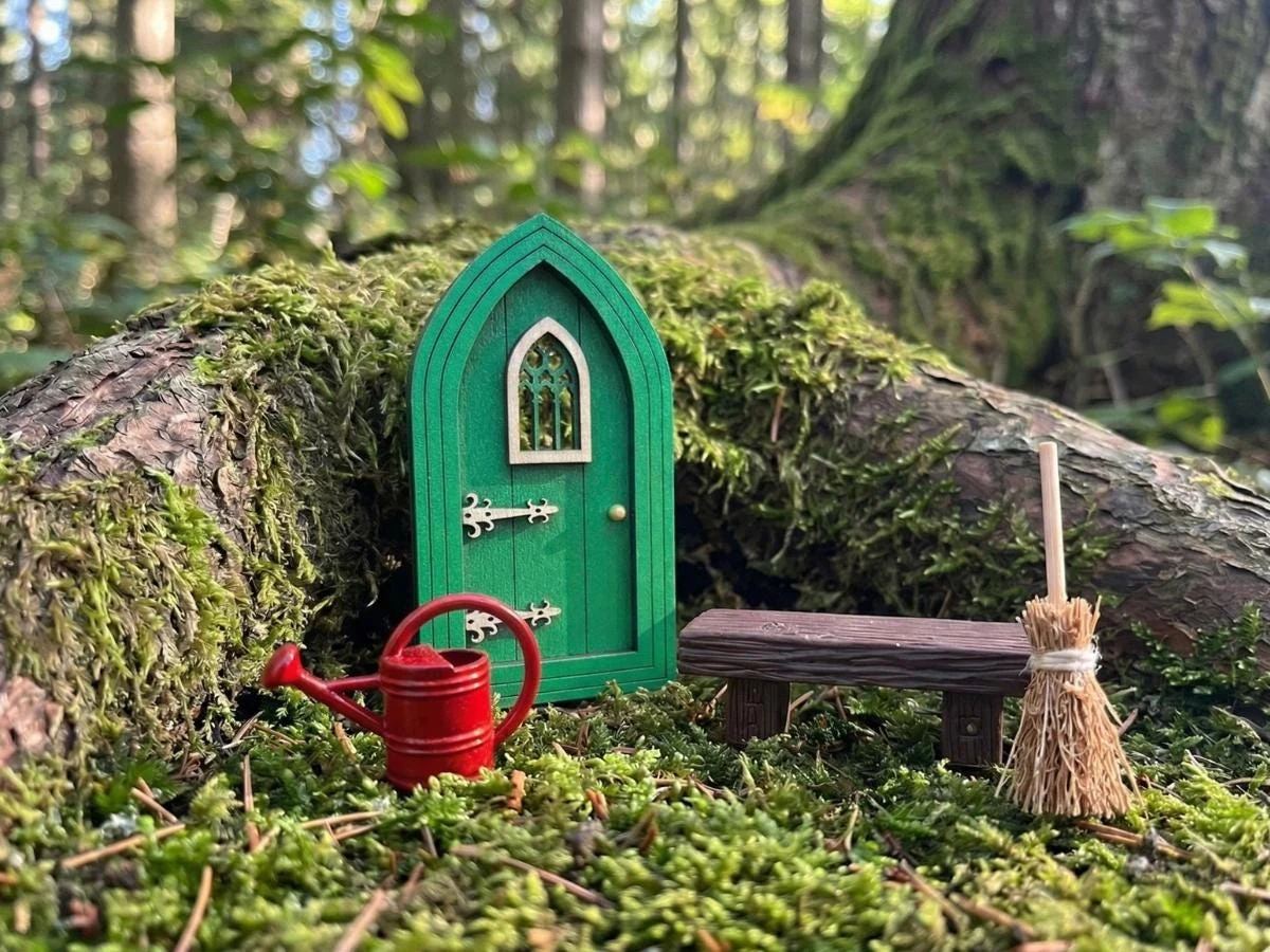 A real green Mary fairy door nestled by a tree, with a tiny bench, broom and watering can
